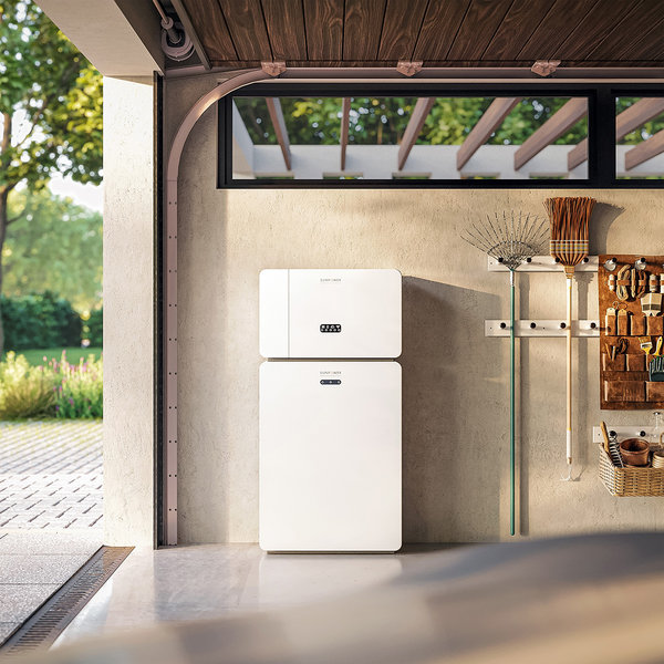 Un moderno sistema di accumulo fotovoltaico a colonna, completamente bianco, installato su una parete di cemento grezzo in un garage luminoso, con attrezzi da giardino appesi a destra e una vista sull'esterno verde a sinistra.
